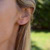 Woman wearing a Birthstone Stud Earring featuring a 4 mm briolette cut Amethyst bezel set in 14k yellow gold