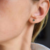 Woman wearing a Birthstone Stud Earring featuring a 4 mm briolette cut Emerald bezel set in 14k yellow gold