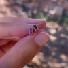 Close-up of a woman's fingers holding two Providence Ruby stud earrings with petite baguette stones set in 14k yellow gold