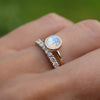Hand wearing two rings including a 1.6 mm wide 14k gold Grand ring featuring one 6 mm briolette cut bezel set moonstone