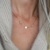 Woman wearing a 14k gold Classic necklace featuring two birthstones and one 1/4” flat disc engraved with a heart symbol
