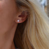 Woman wearing a 14k yellow gold Grand stud earring featuring one 6 mm briolette cut bezel set pink opal