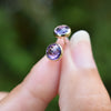 Fingers holding a pair of 14k yellow gold Grand stud earrings each featuring one 6 mm briolette cut bezel set amethyst