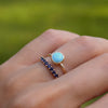 Hand wearing two rings including a 1.6 mm wide 14k gold Grand ring featuring one 6 mm briolette cut bezel set turquoise
